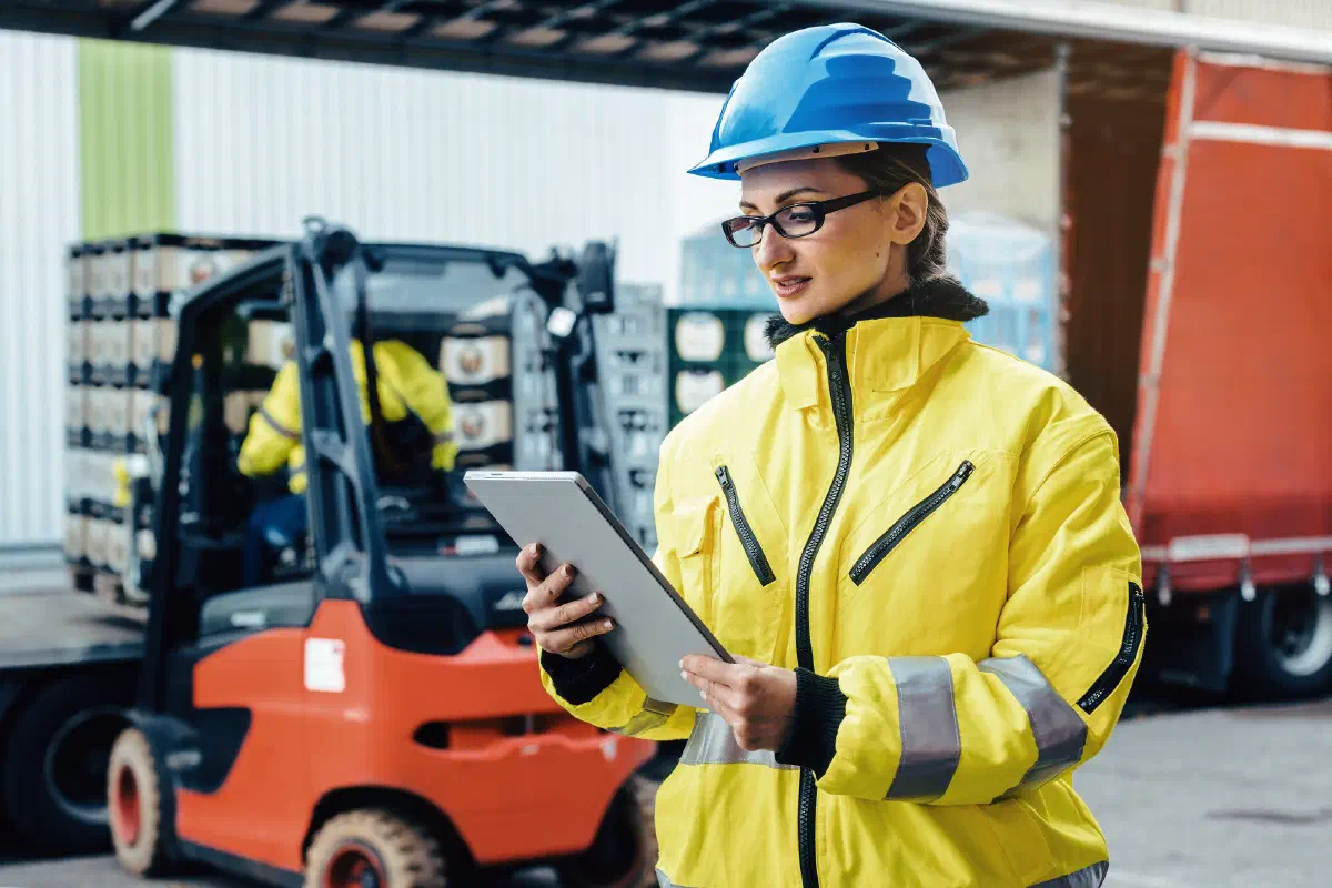 A logistics worker in a hard hat and hi-vis jacket checks a tablet at a loading dock beside a forklift